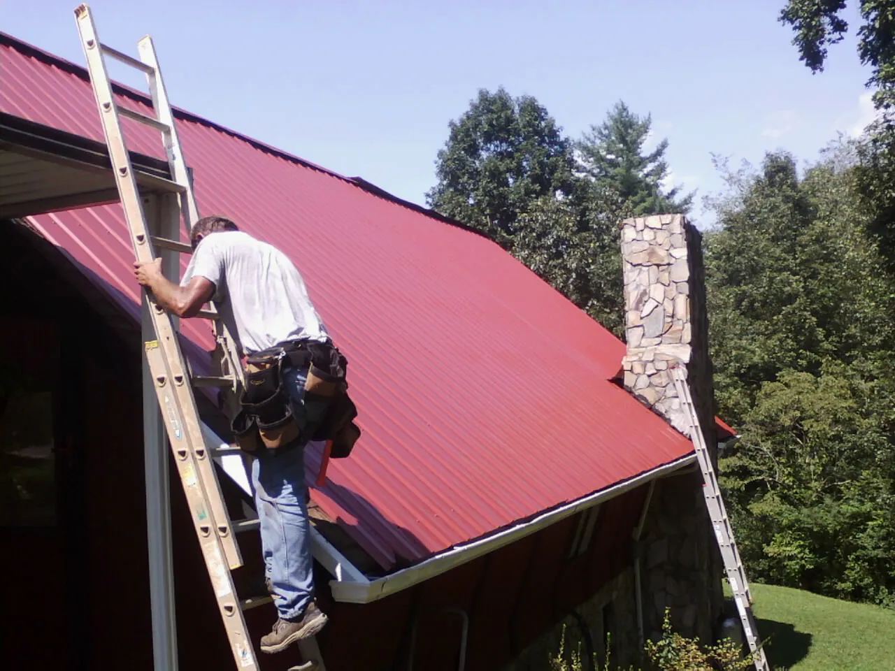 Roofer on a ladder beside a red metal roof and stone chimney — Soffit & Fascia Repair by Roofing Brothers in Blacklick